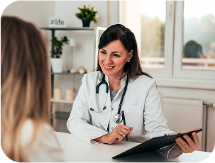 A healthcare professional wearing a white coat and stethoscope is seated at a desk, holding a clipboard and pen while speaking with another person across the table in a bright clinical setting.