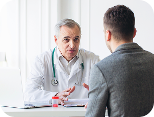 A healthcare professional wearing a white coat with a stethoscope around the neck is seated at a desk, speaking with another person in a bright clinical setting.
